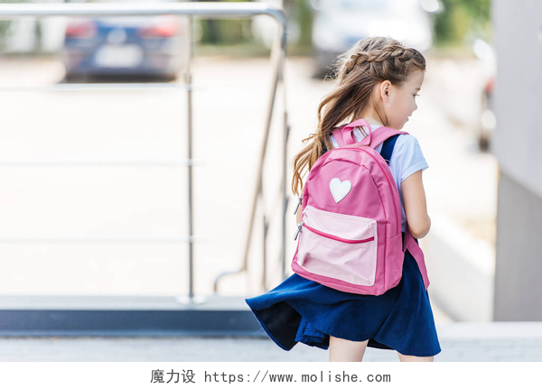 一个穿着蓝色的裙子小女孩去上学小女孩的后方看法与背包在街道